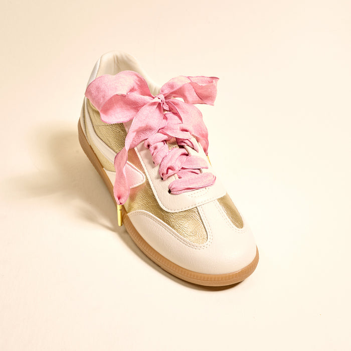 A white and gold sneaker with a gum sole is displayed on a light beige background, featuring The Darling Effect's Frilly Pink Step It Up Shoelaces—wide, crinkled pink satin laces tied in a bow for a unique and stylish look.