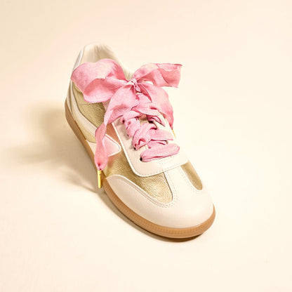 A white and gold sneaker with a gum sole is displayed on a light beige background, featuring The Darling Effect's Frilly Pink Step It Up Shoelaces—wide, crinkled pink satin laces tied in a bow for a unique and stylish look.