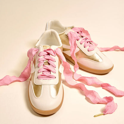 A pair of white and gold sneakers with light brown soles is displayed on a light background, featuring The Darling Effect's Frilly Pink Step It Up Shoelaces.