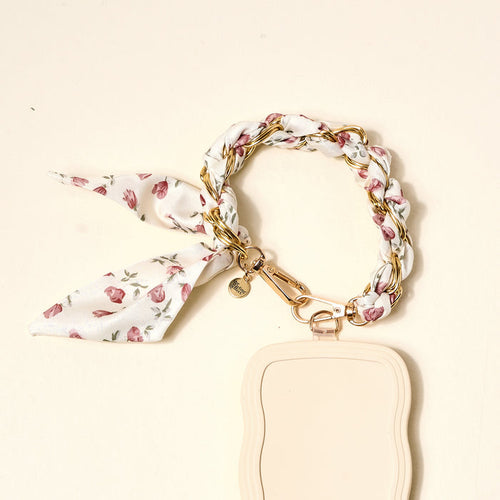 The Darling Effect’s Garden Party Call Me Later Phone Scarf features a gold chain bracelet entwined with a white floral scarf and gold clasp, attached to a beige keychain—a chic essential for your accessories.