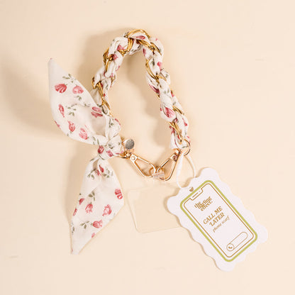 The Darling Effect’s Garden Party Call Me Later Phone Scarf is a floral keychain with a braided strap, gold clasp, and bow. On a light background, it displays a scalloped tag reading CALL ME with a phone graphic.