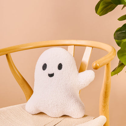 The Darling Effect Ghost Throw Pillow, a sherpa pillow shaped like a smiling white ghost with black eyes and mouth, sits on a light wooden chair by a green leafy plant—perfect for adding spooky-cute Halloween decor to any room.