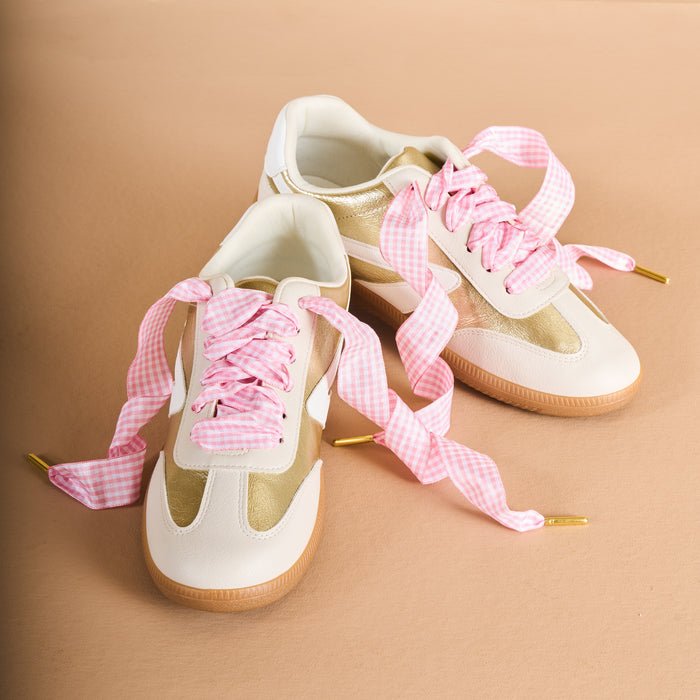Two beige and white sneakers with tan soles are shown on a tan background, laced with The Darling Effect’s Gingham Pink Step It Up Shoelaces—wide pink and white gingham laces with gold tips for a playful yet polished look.
