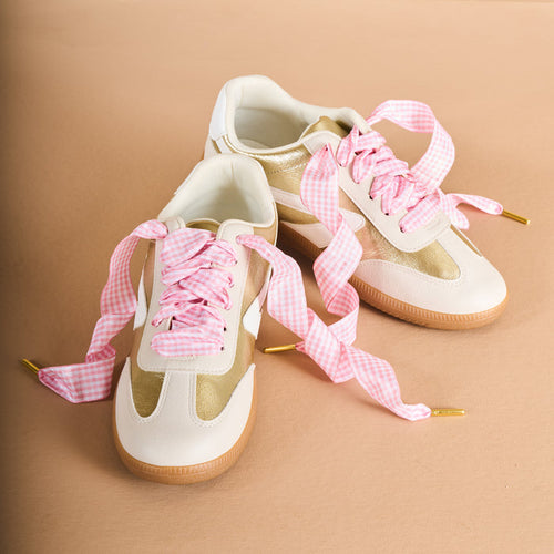 Two beige and white sneakers with tan soles are shown on a tan background, laced with The Darling Effect’s Gingham Pink Step It Up Shoelaces—wide pink and white gingham laces with gold tips for a playful yet polished look.