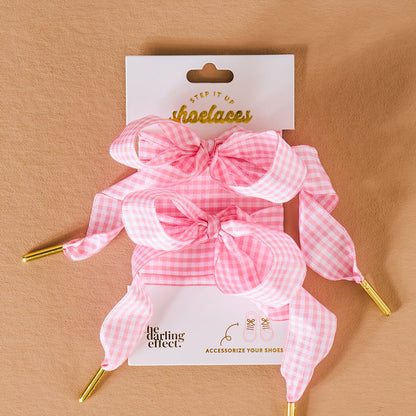 A pair of pink and white gingham durable shoelaces with gold tips, styled in bows and displayed on a branded card reading Step it up shoelaces from The Darling Effect, set against a tan background.