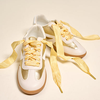 A pair of white sneakers with metallic gold accents and The Darling Effect's Gingham Yellow Step It Up Shoelaces are placed on a light beige surface.