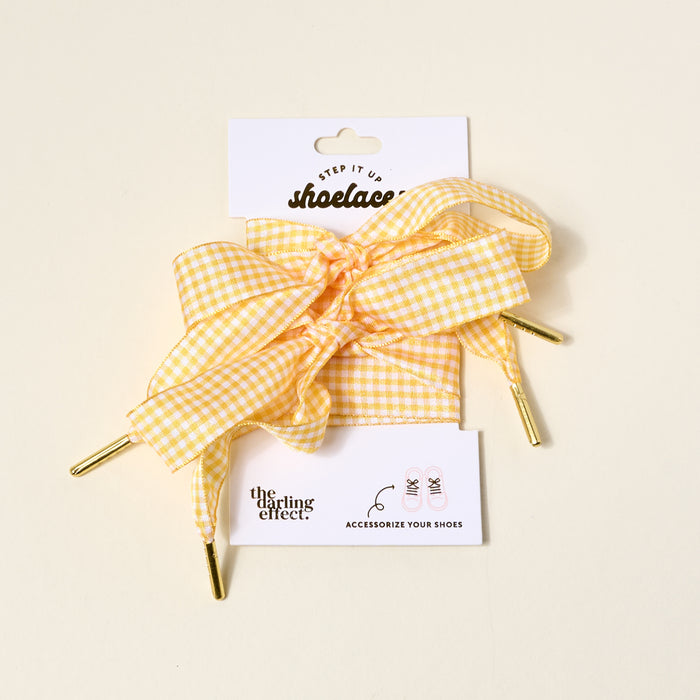 Gingham Yellow Step It Up Shoelaces by The Darling Effect feature yellow and white gingham with gold aglets, tied in a bow on packaging labeled with the brand name and product, plus a sneaker illustration at the bottom.