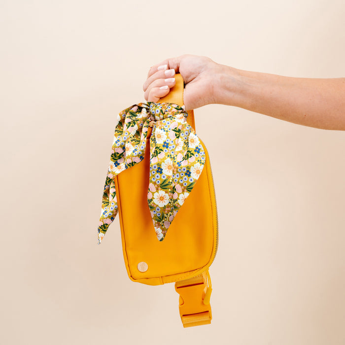 A hand holds The Darling Effect's Golden Glow Belt Bag—hands-free convenience—with a floral-patterned scarf tied in a bow on the handle, set against a plain light beige background.