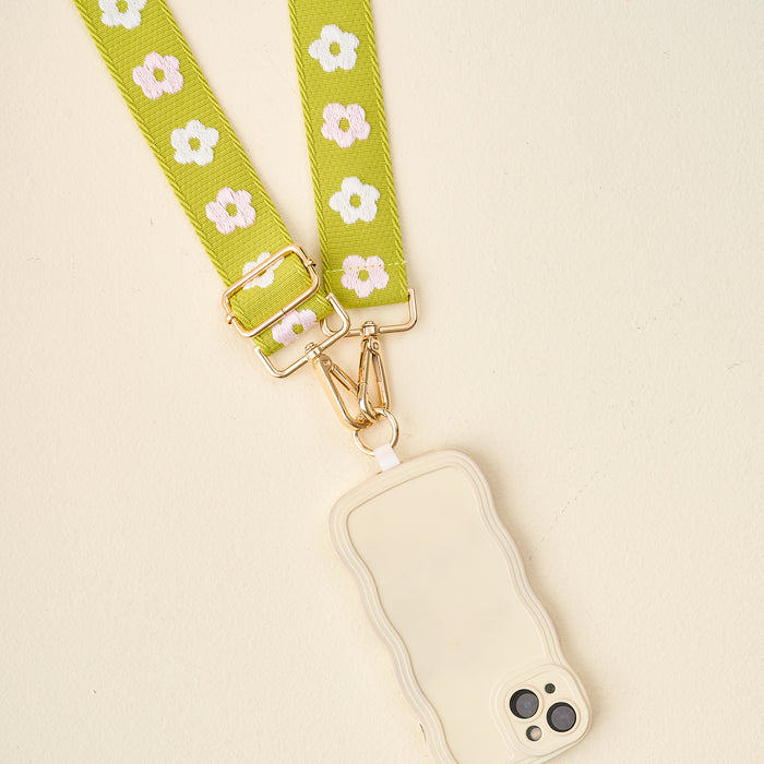 The Darling Effect’s Green Hold the Phone Crossbody Strap, with a white and yellow floral pattern and gold hardware, is paired with a cream-colored wavy-edge phone case on a light background.