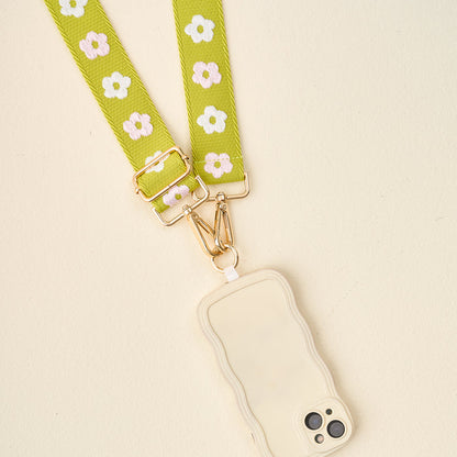 The Darling Effect’s Green Hold the Phone Crossbody Strap, with a white and yellow floral pattern and gold hardware, is paired with a cream-colored wavy-edge phone case on a light background.