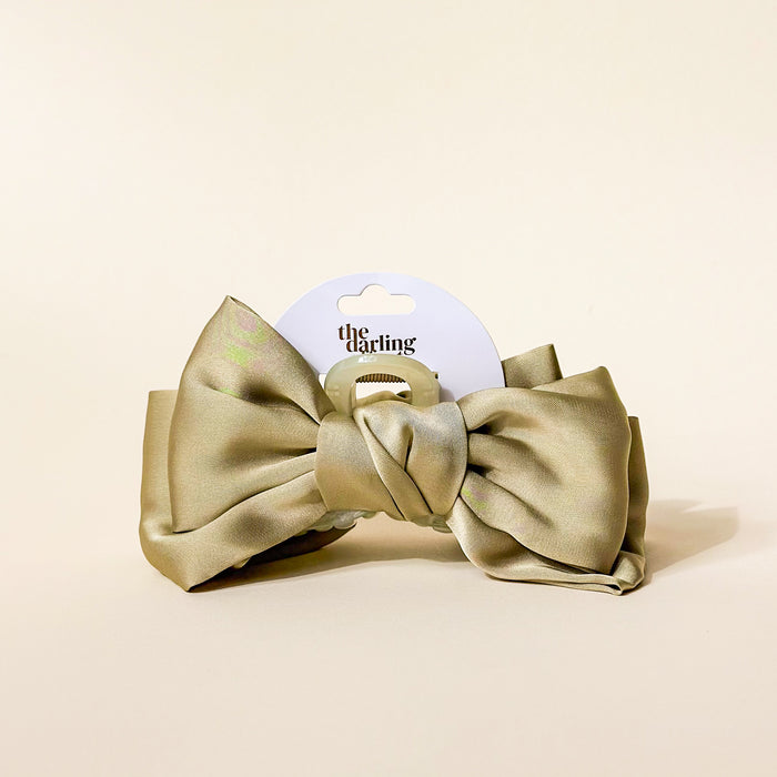 The Darling Effect Green Satin Bow Claw Clip, a large light olive green hair accessory with a central knot, is displayed upright on beige packaging labeled "the darling." Perfect for updos.