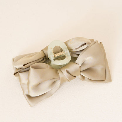 The Darling Effect’s Green Satin Bow Claw Clip secures a beige satin bow, creating an elegant updo accessory. Displayed on a light, textured surface, the translucent clip adds a soft green accent to your hairstyle.