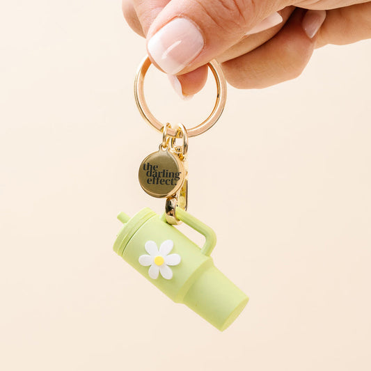 Hand holding The Darling Effect Green Tiny Tumbler Keychain featuring a white daisy, handle, straw, and a round branded tag. Also includes a gold flower keychain charm.