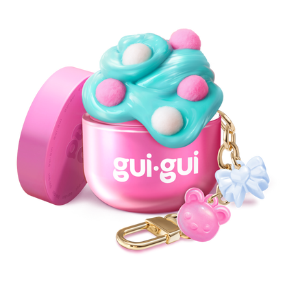 The Gui Gui Mini Backpack Charm Slime by Moose Toys is a small pink container with blue and pink fluffy slime, white and pink dots, plus a gold keychain featuring a pink bear charm and a blue bow charm.