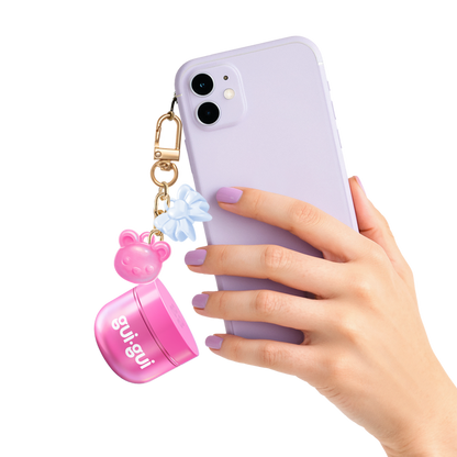 A hand with pastel purple nails holds a lavender smartphone, adorned with a keychain featuring the Moose Toys Gui Gui Mini Backpack Charm Slime, along with a pink bear-shaped charm and blue bow.