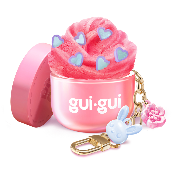 The Gui Gui Mini Backpack Charm Slime by Moose Toys features pink slime with blue heart shapes in a pink gui.gui-labeled container, topped with a gold keychain, blue bunny charm, and pink flower. The lid rests beside it.