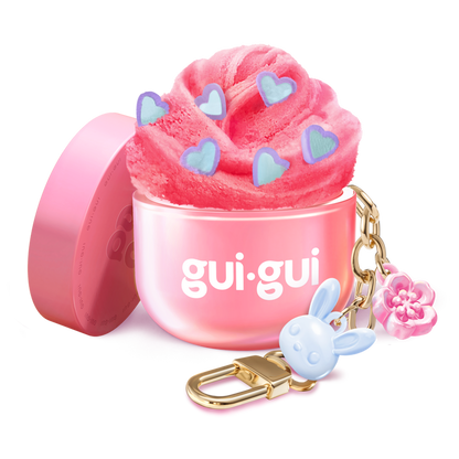 The Gui Gui Mini Backpack Charm Slime by Moose Toys features pink slime with blue heart shapes in a pink gui.gui-labeled container, topped with a gold keychain, blue bunny charm, and pink flower. The lid rests beside it.