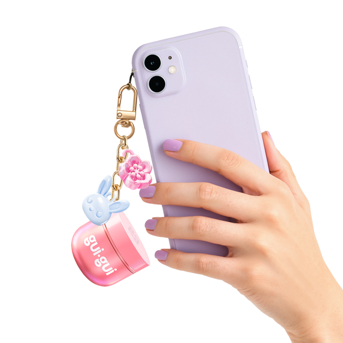 A hand with lavender-painted nails holds a lavender smartphone with a keychain featuring a pink flower, a blue bunny charm, and the Moose Toys Gui Gui Mini Backpack Charm Slime.
