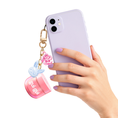 A hand with lavender-painted nails holds a lavender smartphone with a keychain featuring a pink flower, a blue bunny charm, and the Moose Toys Gui Gui Mini Backpack Charm Slime.