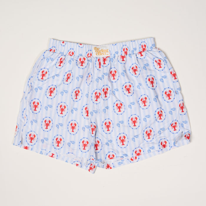 The Darling Effect’s Harbor House All Day Boxer Shorts feature light blue and white stripes with red lobsters and blue lifebuoys, a comfy elastic waistband, breathable fabric, and a signature label—ideal for trendy loungewear.