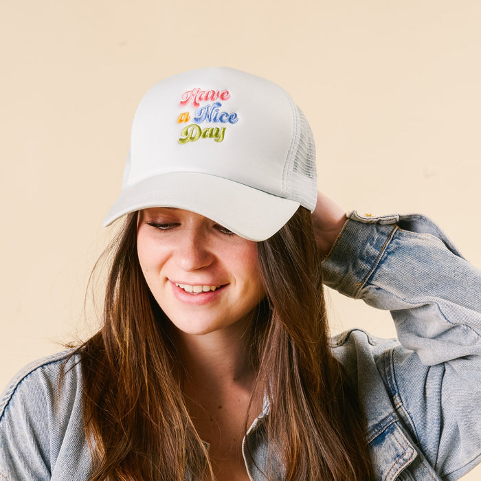 A smiling woman wears the-darling-effect's Have a Nice Day Trucker Hat, featuring colorful retro script embroidery, paired with a denim jacket.