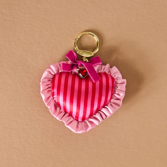 The Darling Effect’s Heartthrob Puffy Heart Bag Charm—a pink heart-shaped keychain with red stripes, ruffled satin trim, and a pink bow on a gold key ring—makes an adorable Valentine’s Day purse accessory, shown on a beige surface.