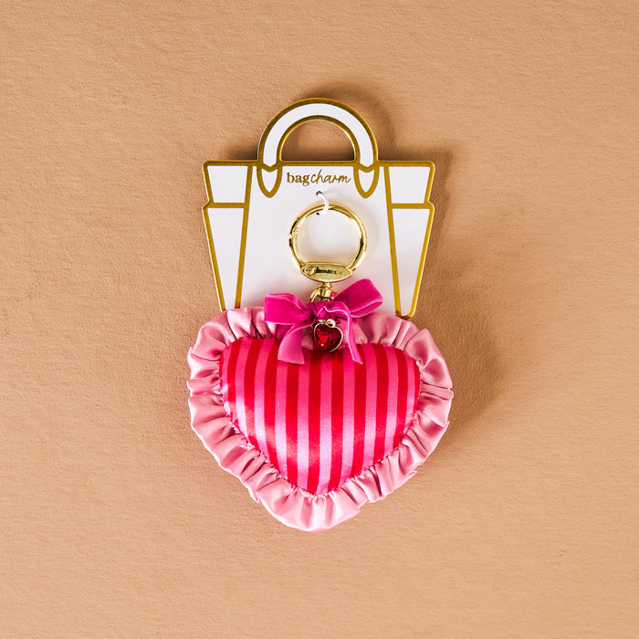 The Darling Effect's Heartthrob Puffy Heart Bag Charm features pink stripes, ruffled edges, a small pink bow, and gold hardware—an adorable Valentine’s Day accessory shown on a beige background.