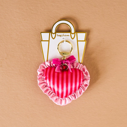 The Darling Effect's Heartthrob Puffy Heart Bag Charm features pink stripes, ruffled edges, a small pink bow, and gold hardware—an adorable Valentine’s Day accessory shown on a beige background.