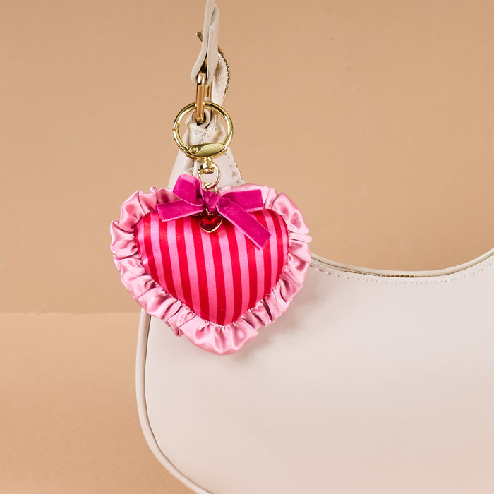 A close-up of a white handbag featuring The Darling Effect’s Heartthrob Puffy Heart Bag Charm—pink, striped, heart-shaped with ruffles and a bow—hanging from the handle on a gold clasp, standing out against a plain beige background.