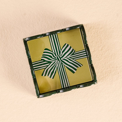 A rectangular gift box with a yellow, green, and white striped lid and bow displays The Darling Effect’s Holiday Gift Bow Green Scallop Trinket Tray, perfect for gifting, set on a light beige textured surface.