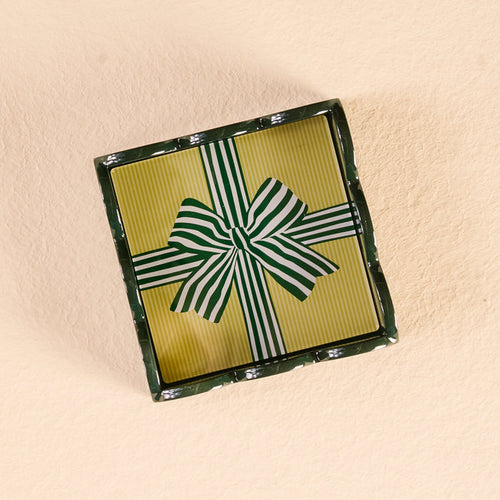 A rectangular gift box with a yellow, green, and white striped lid and bow displays The Darling Effect’s Holiday Gift Bow Green Scallop Trinket Tray, perfect for gifting, set on a light beige textured surface.