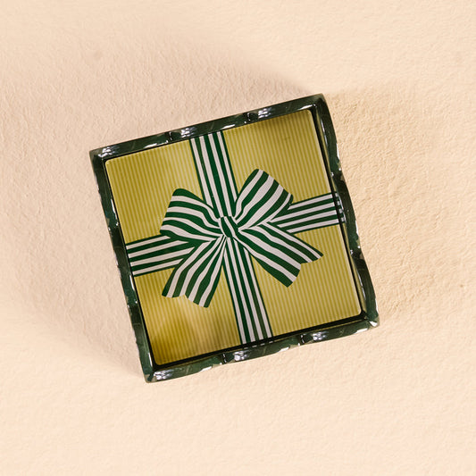 A rectangular gift box with a yellow, green, and white striped lid and bow displays The Darling Effect’s Holiday Gift Bow Green Scallop Trinket Tray, perfect for gifting, set on a light beige textured surface.