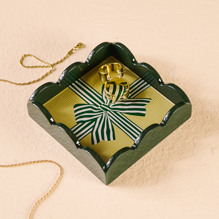 A gold necklace and ring rest in The Darling Effect's Holiday Gift Bow Green Scallop Trinket Tray, featuring a striped ribbon design—perfect for stylishly organizing or displaying jewelry on any light, textured surface.