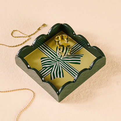 A gold necklace and ring rest in The Darling Effect's Holiday Gift Bow Green Scallop Trinket Tray, featuring a striped ribbon design—perfect for stylishly organizing or displaying jewelry on any light, textured surface.