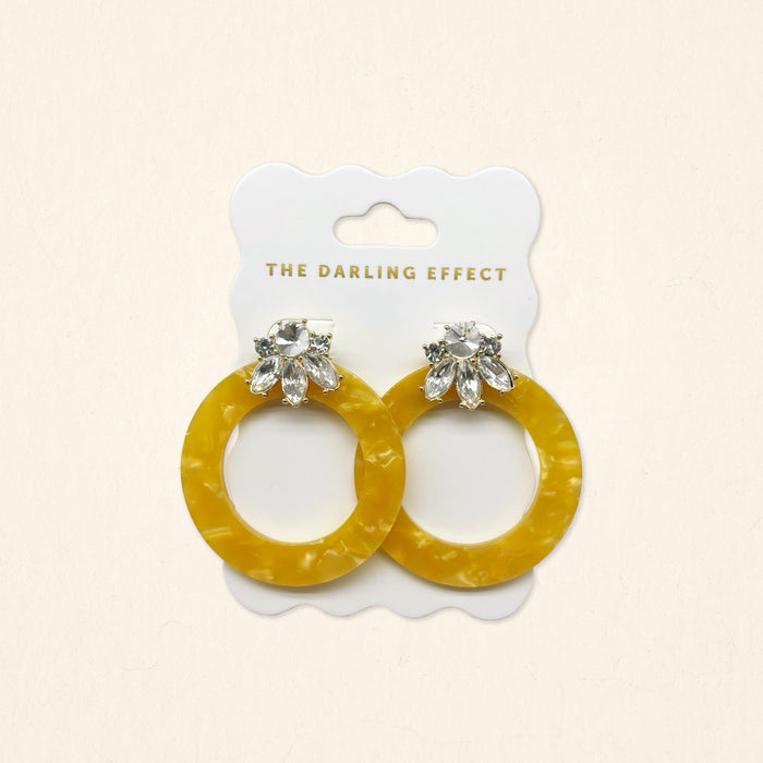 The Darling Effect Honey Gold Glow Getter Earrings feature mustard yellow glossy acetate hoops and crystal rhinestone flower studs on a white scalloped card. Lightweight statement earrings to add a bold, stylish touch to any outfit.