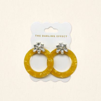 The Darling Effect Honey Gold Glow Getter Earrings feature mustard yellow glossy acetate hoops and crystal rhinestone flower studs on a white scalloped card. Lightweight statement earrings to add a bold, stylish touch to any outfit.