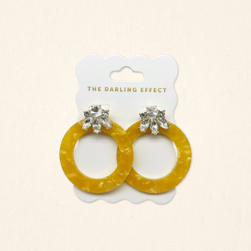 The Darling Effect Honey Gold Glow Getter Earrings feature mustard yellow glossy acetate hoops and crystal rhinestone flower studs on a white scalloped card. Lightweight statement earrings to add a bold, stylish touch to any outfit.