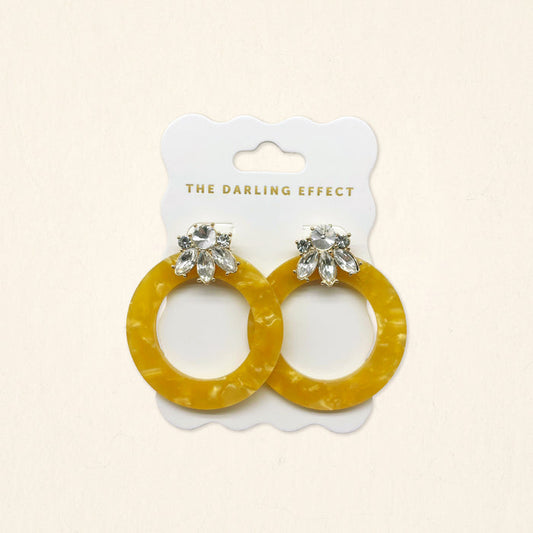 The Darling Effect Honey Gold Glow Getter Earrings feature mustard yellow glossy acetate hoops and crystal rhinestone flower studs on a white scalloped card. Lightweight statement earrings to add a bold, stylish touch to any outfit.