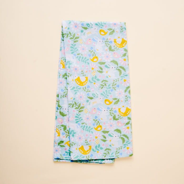 The Hoppy Days Tea Towel by The Darling Effect features light blue fabric adorned with yellow birds, green leaves, and pastel flowers, shown folded on a beige background.