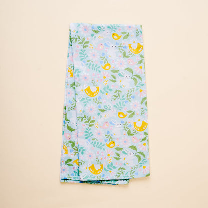 The Hoppy Days Tea Towel by The Darling Effect features light blue fabric adorned with yellow birds, green leaves, and pastel flowers, shown folded on a beige background.