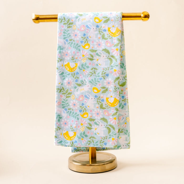 The Darling Effect's Hoppy Days Tea Towel, featuring a whimsical floral and bird print in pastel blue, hangs neatly on a gold metal stand against a light beige background.