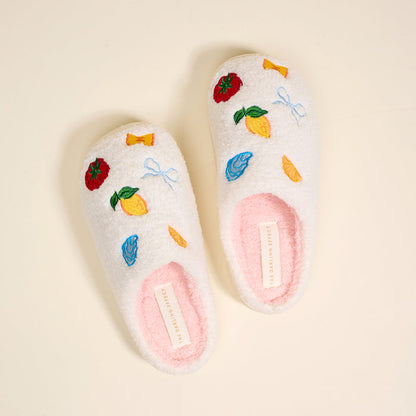 The Darling Effect’s Italian Summer Fuzzy Slippers, shown on a light beige background, are white with a soft pink insole. Embroidered lemons, candy, bows, and strawberries add playful charm along with a visible label.