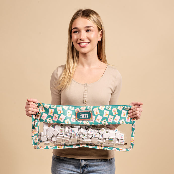 A person wearing a beige top and blue jeans smiles while holding The Darling Effect's It's Mahj Time Teal Mahjong Tile Keeper Pouch filled with mahjong tiles against a neutral background.
