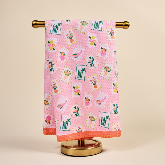 The Darling Effect’s “It’s Mahj Time” Waffle Tea Towel, a pink and absorbent kitchen towel, hangs neatly on a gold stand against a beige background.