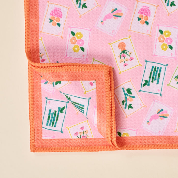 The Darling Effect's It's Mahj Time Waffle Tea Towel features a vibrant pink and orange border with framed flowers, bamboo, and abstract designs. A folded corner shows the same colorful pattern on the absorbent kitchen towel’s underside.