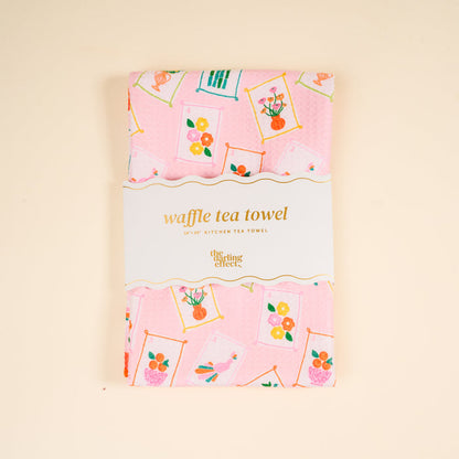 A pink decorative tea towel with framed flower and bird prints, folded and wrapped with a label that reads "It’s Mahj Time Waffle Tea Towel" from The Darling Effect. Absorbent and perfect for everyday kitchen use.