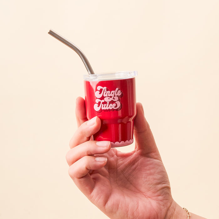 A hand holds the-darling-effect’s Jingle Juice 2 oz Tiny Tumbler, featuring a lid, metal straw, and “Jingle Juice” in white on the front. The background is a plain light beige color.