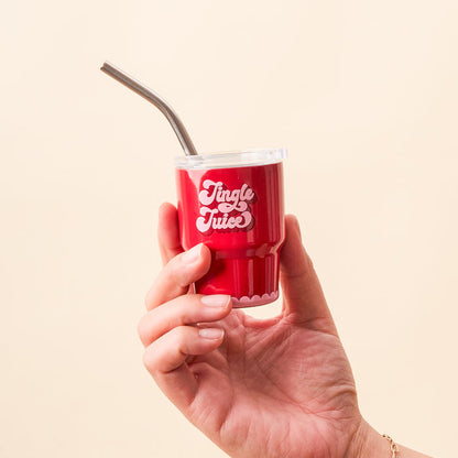 A hand holds the-darling-effect’s Jingle Juice 2 oz Tiny Tumbler, featuring a lid, metal straw, and “Jingle Juice” in white on the front. The background is a plain light beige color.