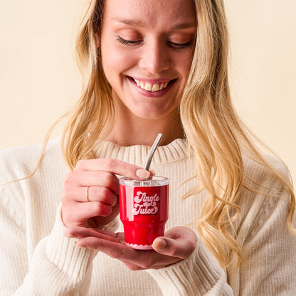 A smiling woman in a cream sweater holds the-darling-effect’s Jingle Juice 2 oz Tiny Tumbler with a metal straw, looking down at her drink against a neutral background.