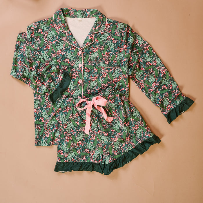 The Darling Effect’s Jolly Sprig Sweet Dreams Pajama Shorts Set features festive holly and berry prints, a soft long-sleeve button-up top, ruffled shorts with a pink ribbon tie, displayed on a light brown background.